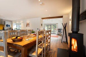 Un coin repas avec une table en bois, des chaises et un panier de fruits. On aperçoit un poêle à bois à l’arrière-plan.