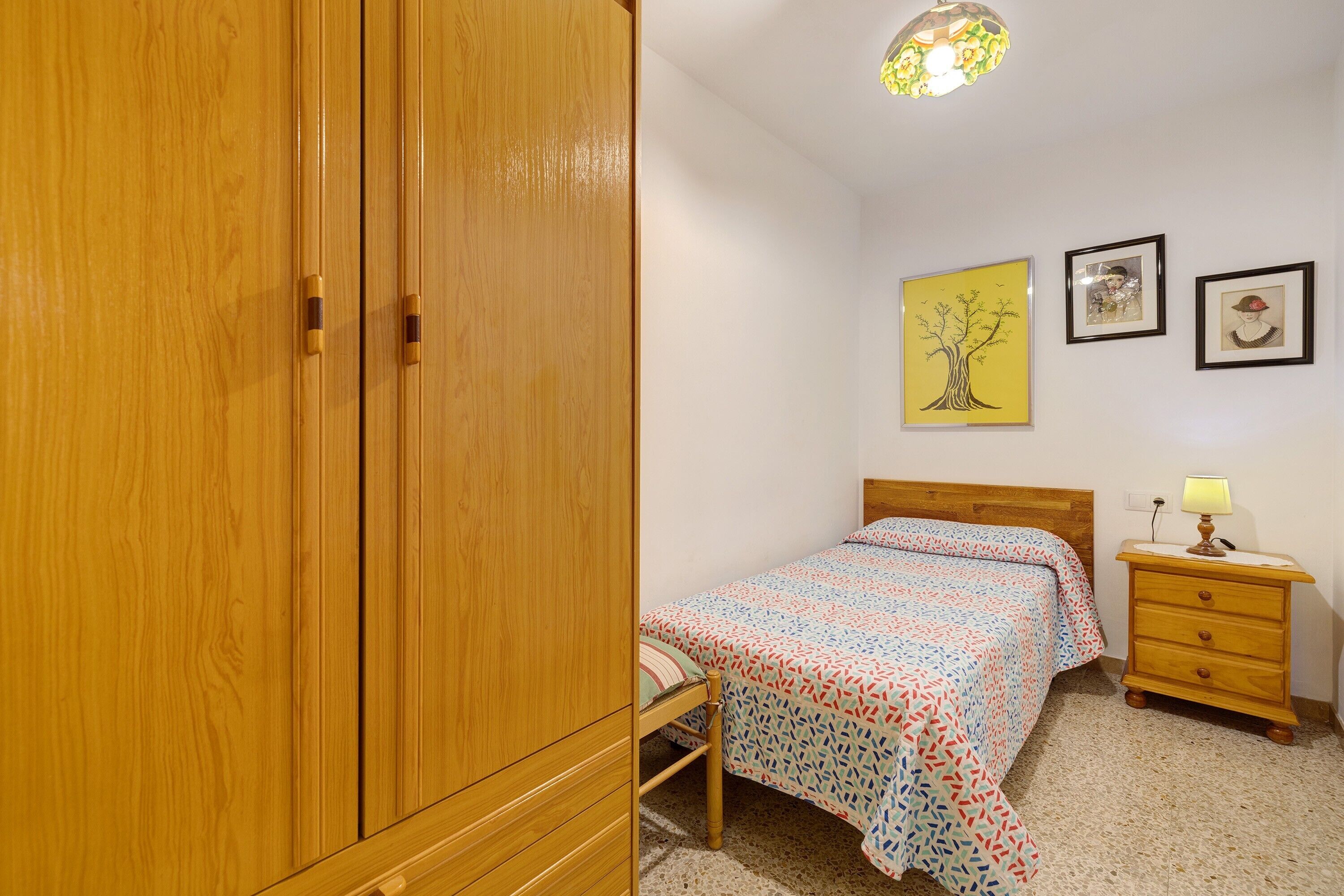 Une chambre avec une armoire en bois, un lit avec une literie à motifs, une table de chevet avec une lampe et des œuvres d’art accrochées au mur.