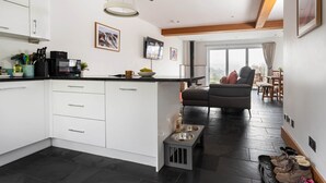 Una cocina moderna con armarios blancos, encimera negra y una zona de comedor con sofá gris y mesa de madera.