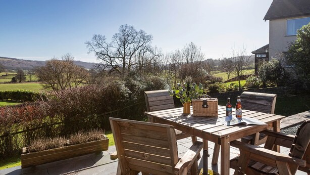 Una mesa de madera al aire libre con sillas, una cesta y botellas, con cielo despejado y colinas al fondo.