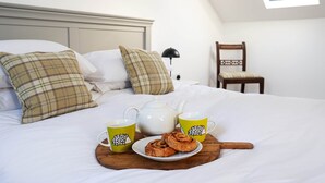 Una cama bien hecha con almohadas, una mesita de noche con lámpara, una silla y una bandeja con tetera, dos tazas y pasteles.