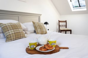 Un lit bien fait, avec des oreillers, une table de chevet sur laquelle se trouve une lampe, une chaise et un plateau avec une théière, deux tasses et des pâtisseries.