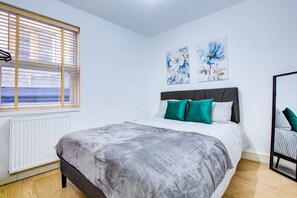 A bedroom with a bed, a radiator, a window with blinds, and two floral wall art pieces. - Perfect Booking Deptford High Street 3 Bed Flat (London)