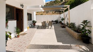 A covered outdoor patio with a table and chairs, potted plants, and a view of a hillside. - Sunny Retreat - Kefalos Stay with Patio & Parking (Kamari)