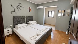 A hotel room with two beds, a nightstand, a wall-mounted air conditioner, and a door leading to another room. - Sunny Retreat - Kefalos Stay with Patio & Parking (Kamari)