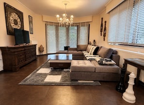 A living room with a sectional sofa, a coffee table, a dark wooden cabinet, a chandelier, and large windows with blinds. - Peaceful Retreat - Kefalos Stay with Patio & Views (Καμάρι)