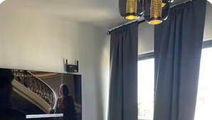 Ein modernes Wohnzimmer mit Flachbildfernseher, weißem Lowboard, einem kleinen Tisch und einem großen Fenster mit Vorhängen.