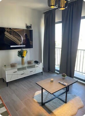 Ein modernes Wohnzimmer mit Flachbildfernseher, weißem Lowboard, einem kleinen Tisch und einem großen Fenster mit Vorhängen.