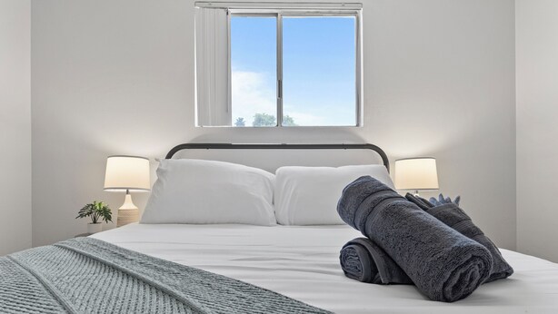 Ein ordentlich bezogenes Bett mit Kissen, ein gerolltes Handtuch und einer Decke. Ein Fenster bietet Blick auf den Himmel und Bäume.