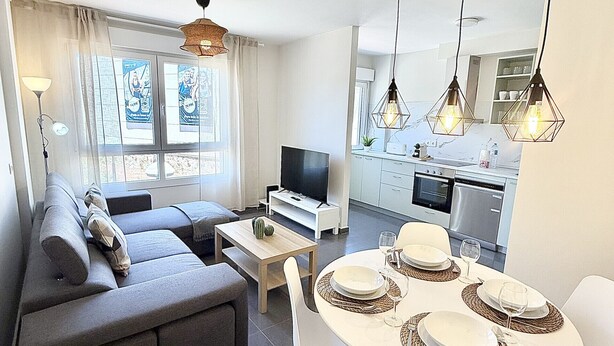 A modern living room with a sofa, TV, dining table with plates and glasses, kitchen area with cabinets and appliances, and pendant lights. - Dragos Suites 105 (Arrecife)