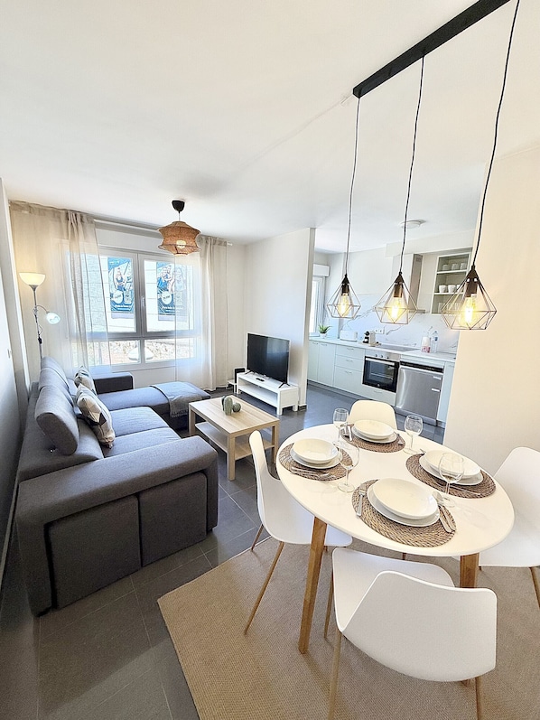 Ein modernes Wohnzimmer mit Sofa, Fernseher, Esstisch mit Tellern und Gläsern, Küchenbereich mit Schränken und Geräten sowie Pendelleuchten.