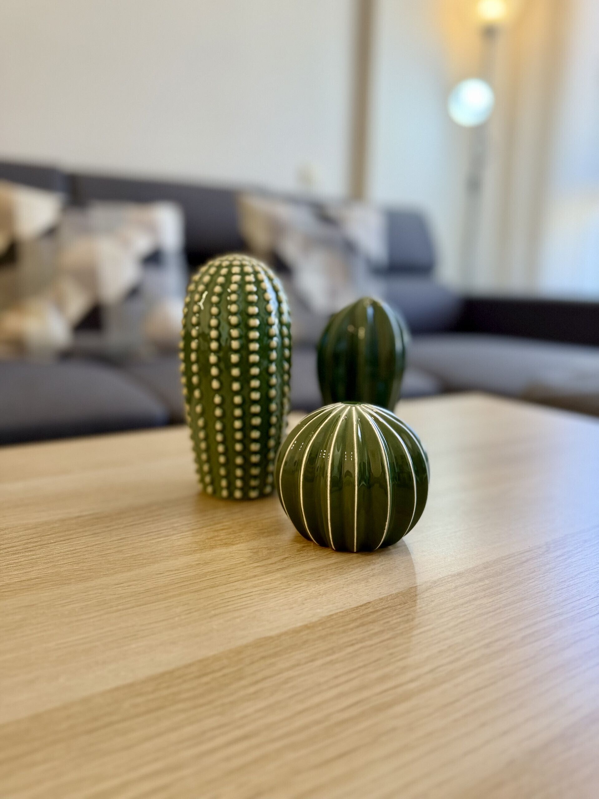 Three green vases on a wooden table in a living room with a sofa and a lamp in the background.