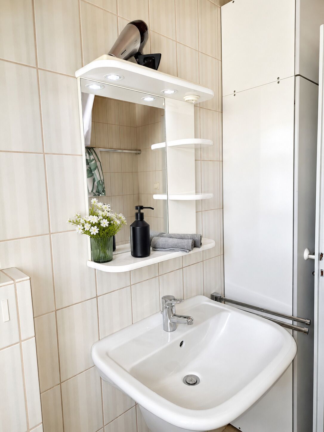 Un baño con un lavamanos blanco, un espejo con iluminación integrada, un perchero y un secador de pelo en un estante.