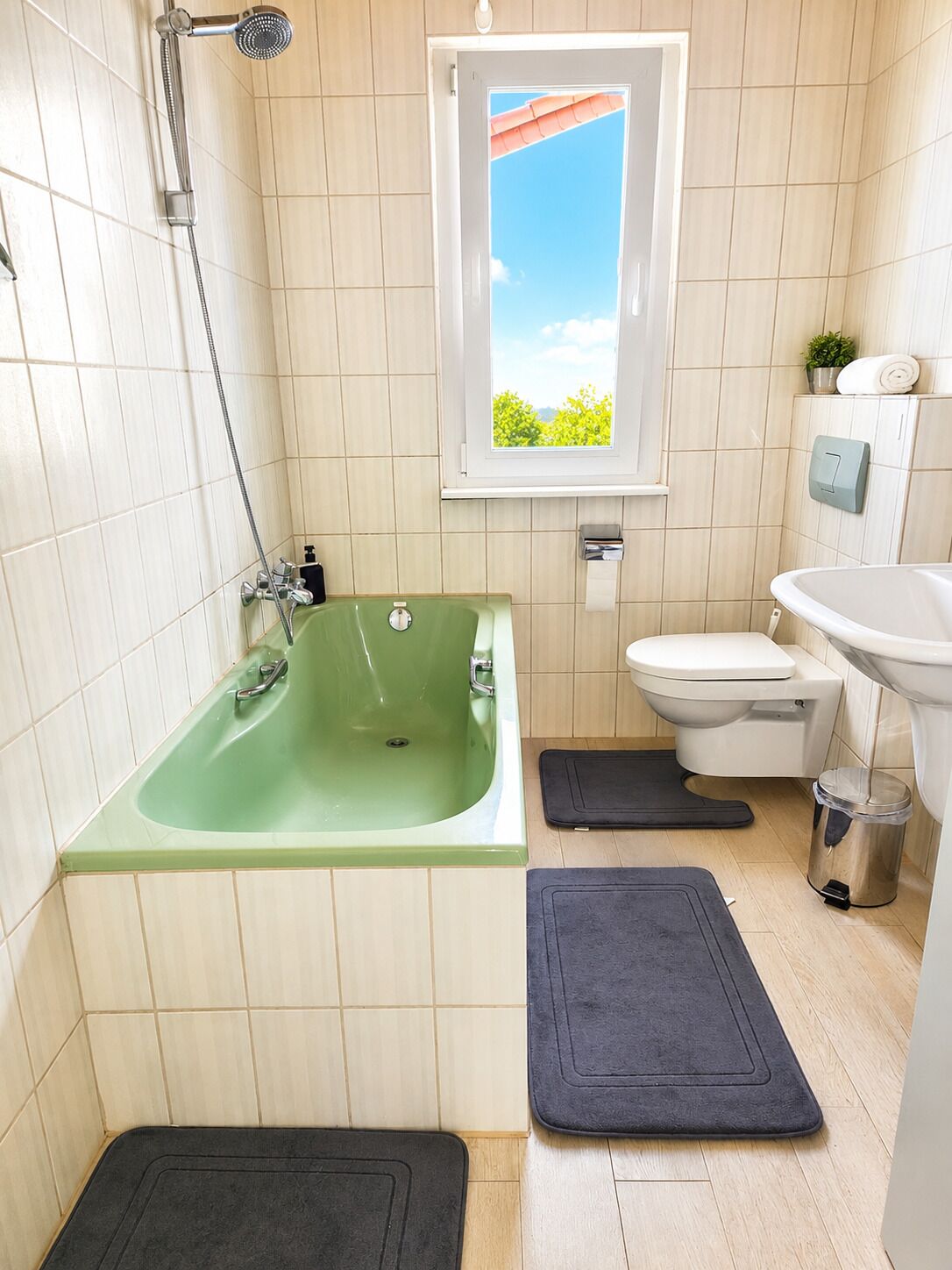 A bathroom with a green bathtub, a toilet, a sink, a window with a view, and two mats on the floor.