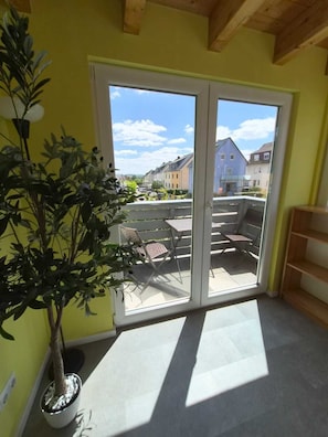 Une pièce dotée d’une porte-fenêtre coulissante donnant sur une terrasse extérieure avec une table et des chaises, un pot de fleurs et une vue sur les maisons voisines.