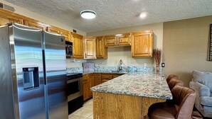 A modern kitchen with a stainless steel refrigerator, wooden cabinets, a granite countertop island, and a bar stool. - Poolside Condo with Bunks - Family Resort Stay at Las Palmas Resort #1625 (St. George)