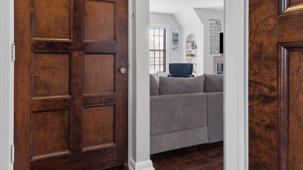 A hallway with a wooden door, a grey sofa, and a white wall with a circular light fixture. - Central-Immaculate-Parking-Fenced Yard-Dining (Fort Wayne)