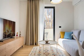 Ein modernes Wohnzimmer mit Flachbildfernseher, rundem Couchtisch, einer Couch mit Kissen, einem gemusterten Teppich und einem Fenster mit Vorhängen.