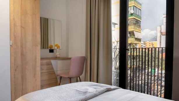 Ein Schlafzimmer mit Bett, Schreibtisch, Stuhl, Spiegel und Blick auf Gebäude durch ein Fenster.