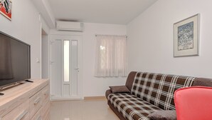 Uma sala de estar com televisão sobre um armário de madeira, um sofá xadrez, uma cadeira vermelha, uma porta e uma janela com cortinas.