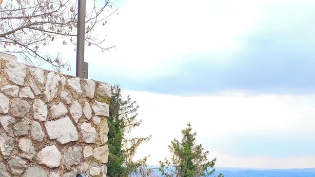A green hammock chair with metal frame, set against a stone wall and metal railing, overlooking a scenic landscape.