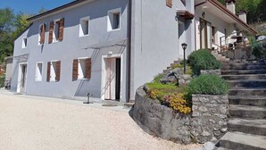 A two-story building with a white facade, multiple windows with shutters, a staircase leading to the entrance, and a small garden area with rocks and plants.