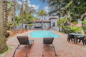 A swimming pool with lounge chairs and tables, surrounded by palm trees and a building with a red roof.