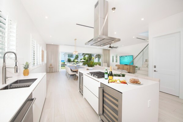 A modern kitchen with a central island, built-in appliances, and a dining area with outdoor views. - Oceans 9, Villa 9: Seaside Lazy River Pool, Sunken BBQ Ocean Lounge + Designer Interiors in West Bay (Grand Cayman)