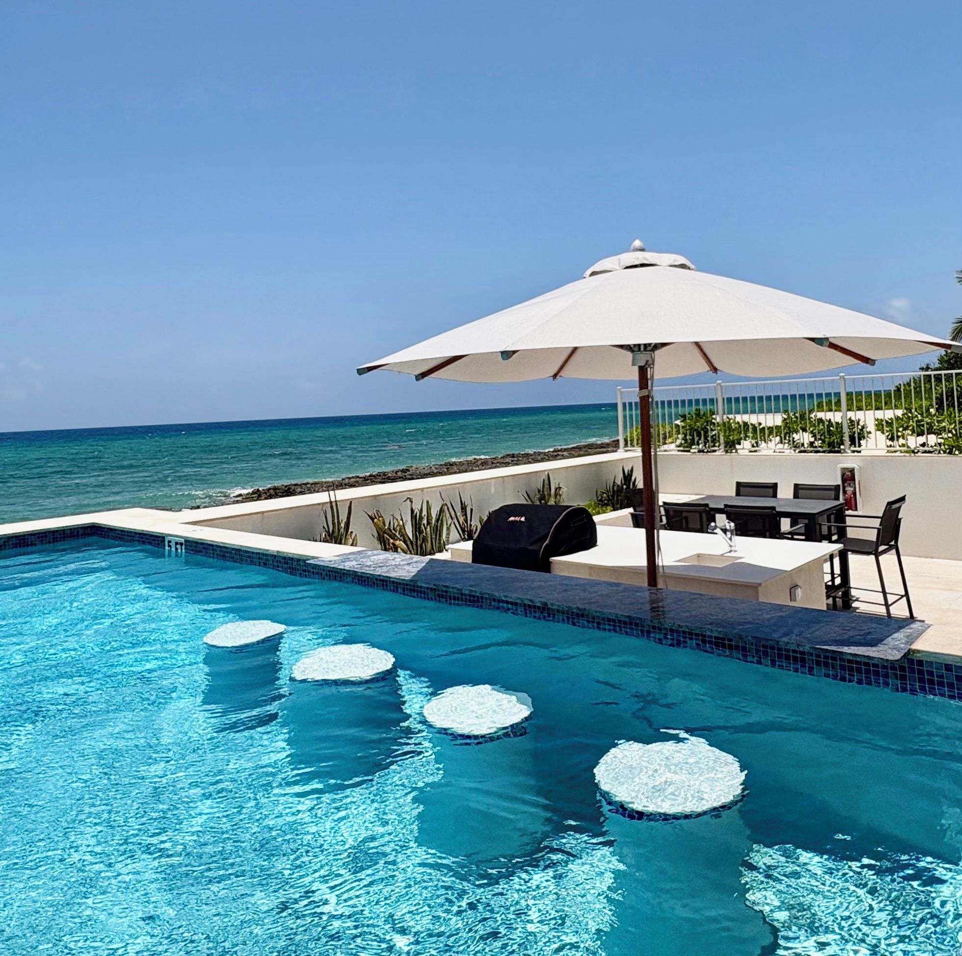 A swimming pool with clear blue water, a white umbrella, and a view of the ocean.