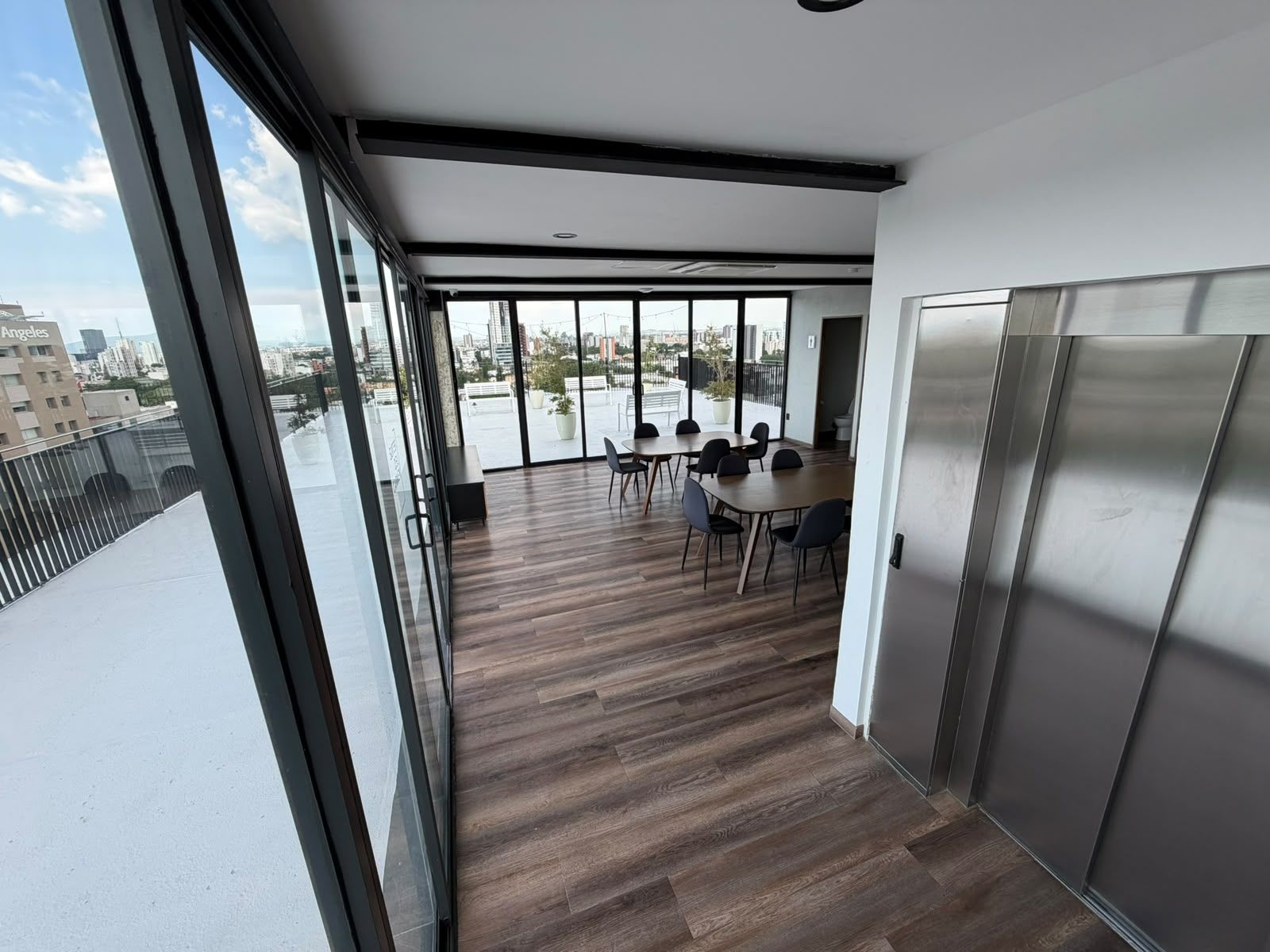 Een modern interieur met een grote glazen deur die uitkomt op een terras. Binnen ligt een houten vloer, staat een tafel met stoelen en is er een groot raam met uitzicht over de stad.