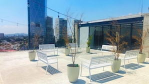A rooftop terrace with white benches and potted plants, overlooking a cityscape with modern buildings. - Tribu Consulado (Guadalajara)