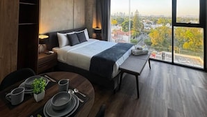 A modern hotel room with a large bed, a dining table with plates and cutlery, a bench, and a view of the city through large windows. - Tribu Consulado (Guadalajara)