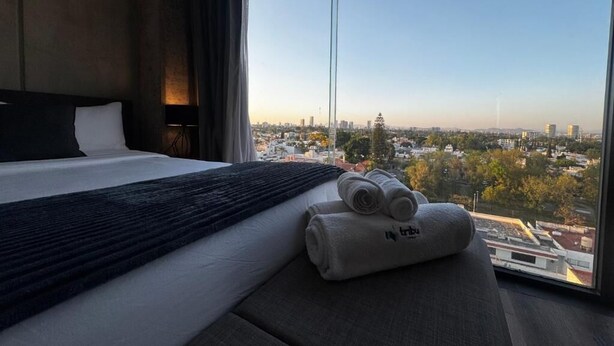 A hotel room with a large bed, a view of the cityscape, and a window with curtains. - Tribu Consulado (Guadalajara)