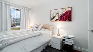 Una camera d'albergo con un letto, un comodino con una lampada, un quadro appeso al muro e una finestra con tende.