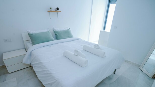 Una habitación de hotel moderna con una cama grande, ropa de cama blanca, una mesita de noche pequeña y una ventana con persianas.