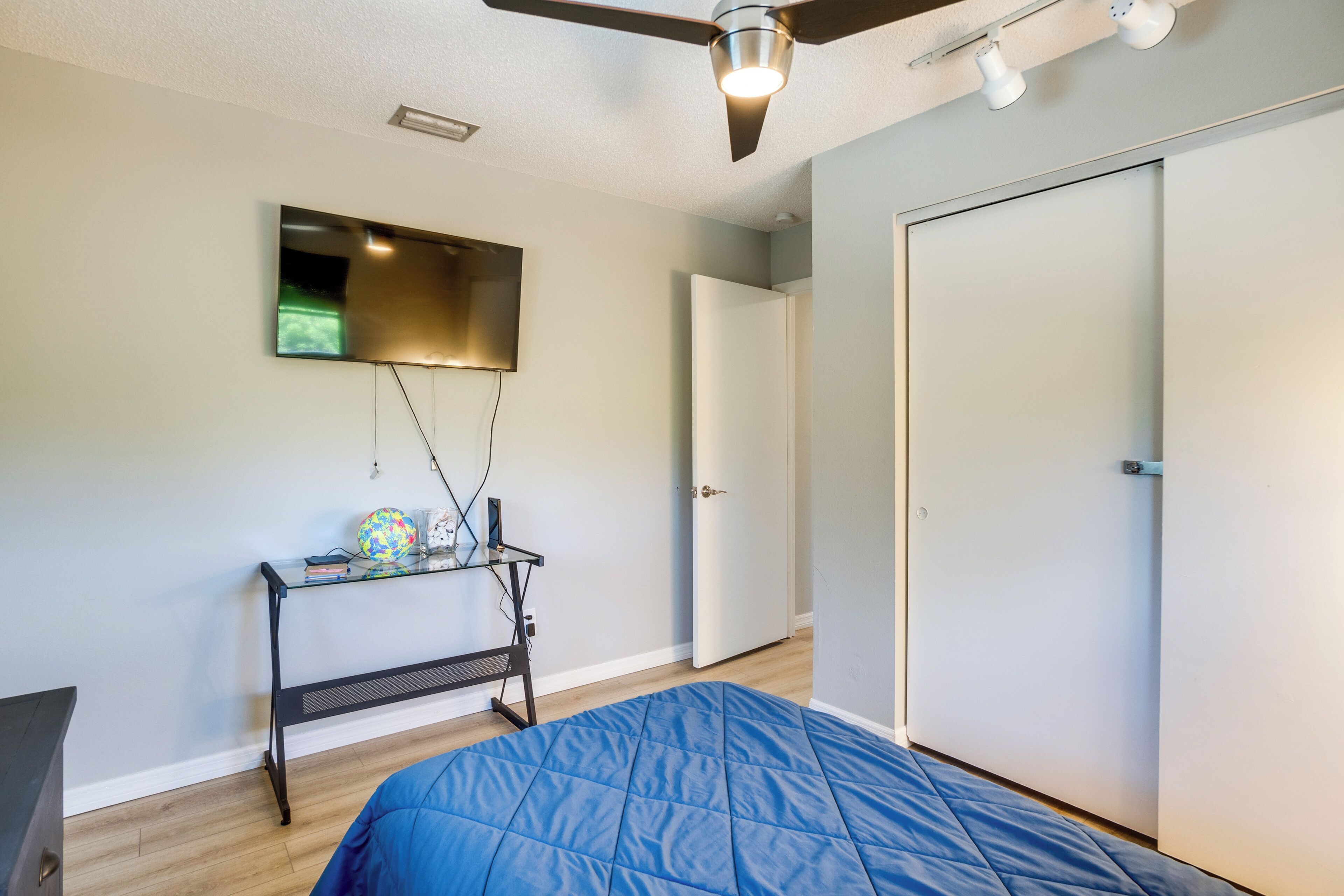 A bedroom with a bed, a ceiling fan, a TV mounted on the wall, a desk with a globe and books, and a door.