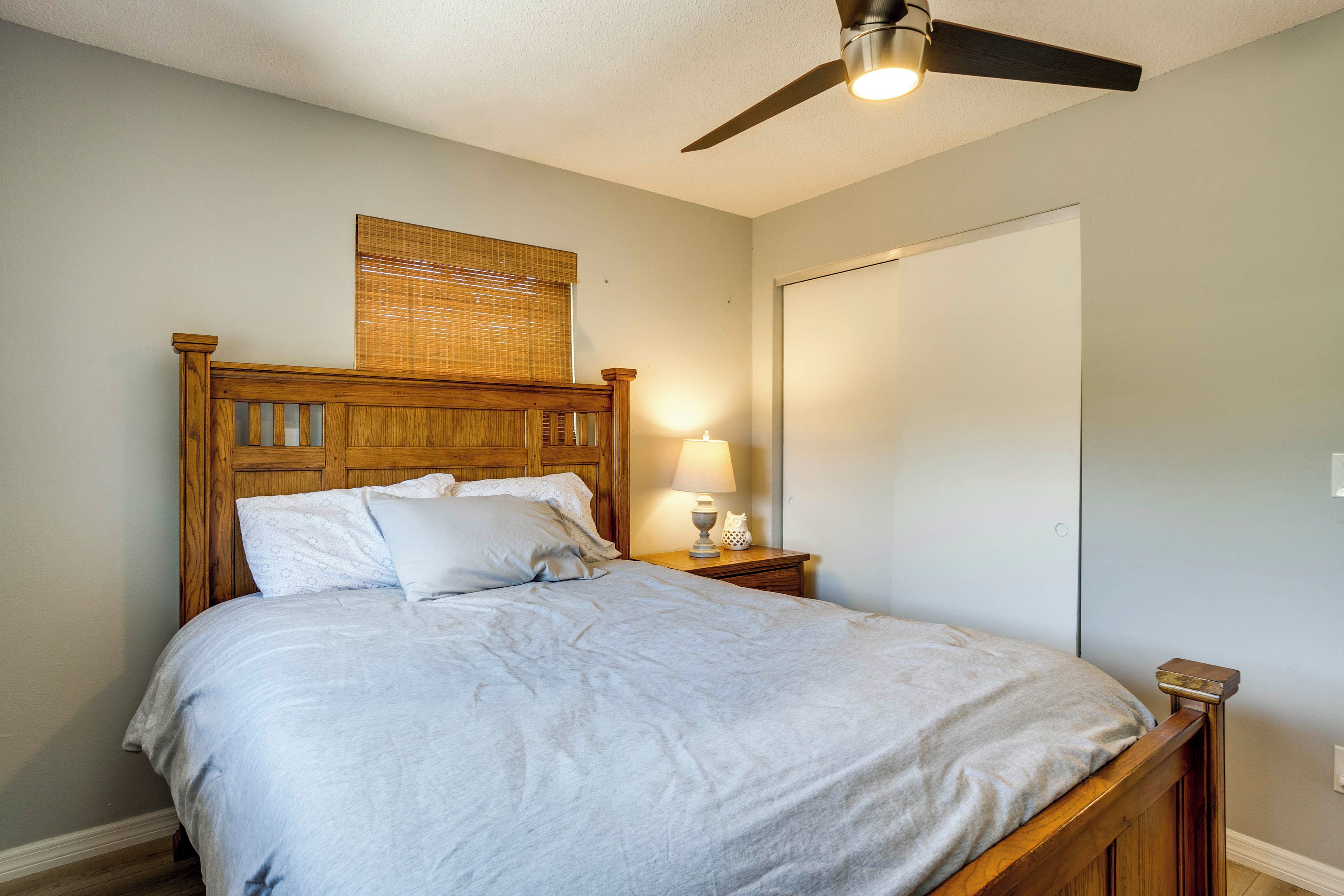 A bedroom with a wooden bed, a nightstand with a lamp, a ceiling fan, and a window with blinds.