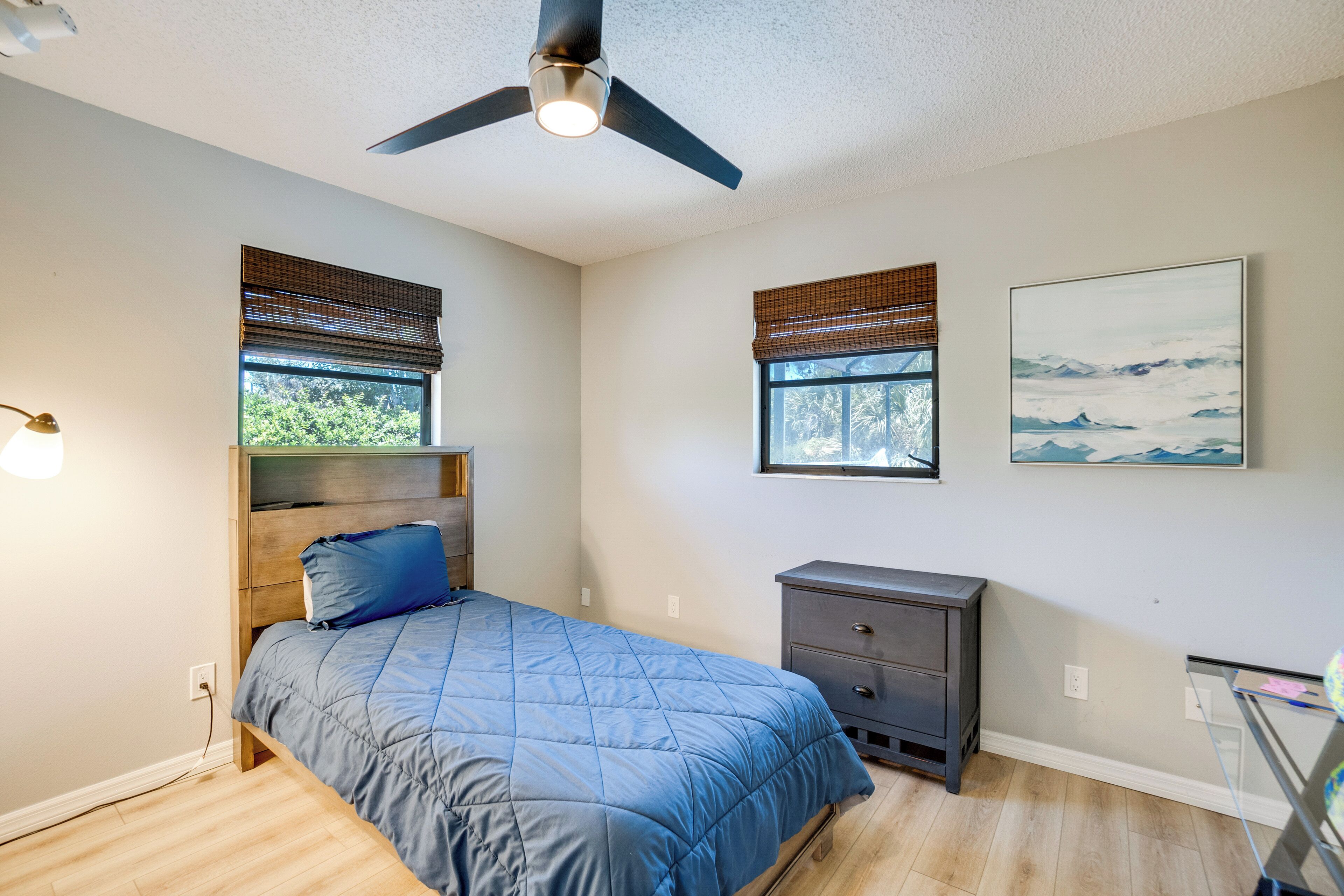 A bedroom with a bed, a ceiling fan, a nightstand, a window with blinds, and a painting on the wall.