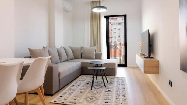 A modern living room with a sofa, a dining table, chairs, a TV, and a patterned rug. - Barrikada-E – Central Apartments with French Balcony & Netflix in Tirana (Tiranë)