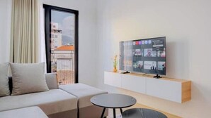A modern living room with a sofa, a small round table, a TV mounted on a white cabinet, and a view of buildings through a large window. - Barrikada-E – Central Apartments with French Balcony & Netflix in Tirana (Tiranë)