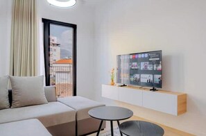 Ein modernes Wohnzimmer mit einer Couch, einem kleinen runden Tisch, einem an einem weißen Schrank montierten Fernseher und Blick auf Gebäude durch ein großes Fenster.