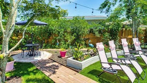 Une terrasse avec une table et des chaises, un petit jardin avec des plantes et une sculpture d’oiseau, et une rangée de chaises longues.