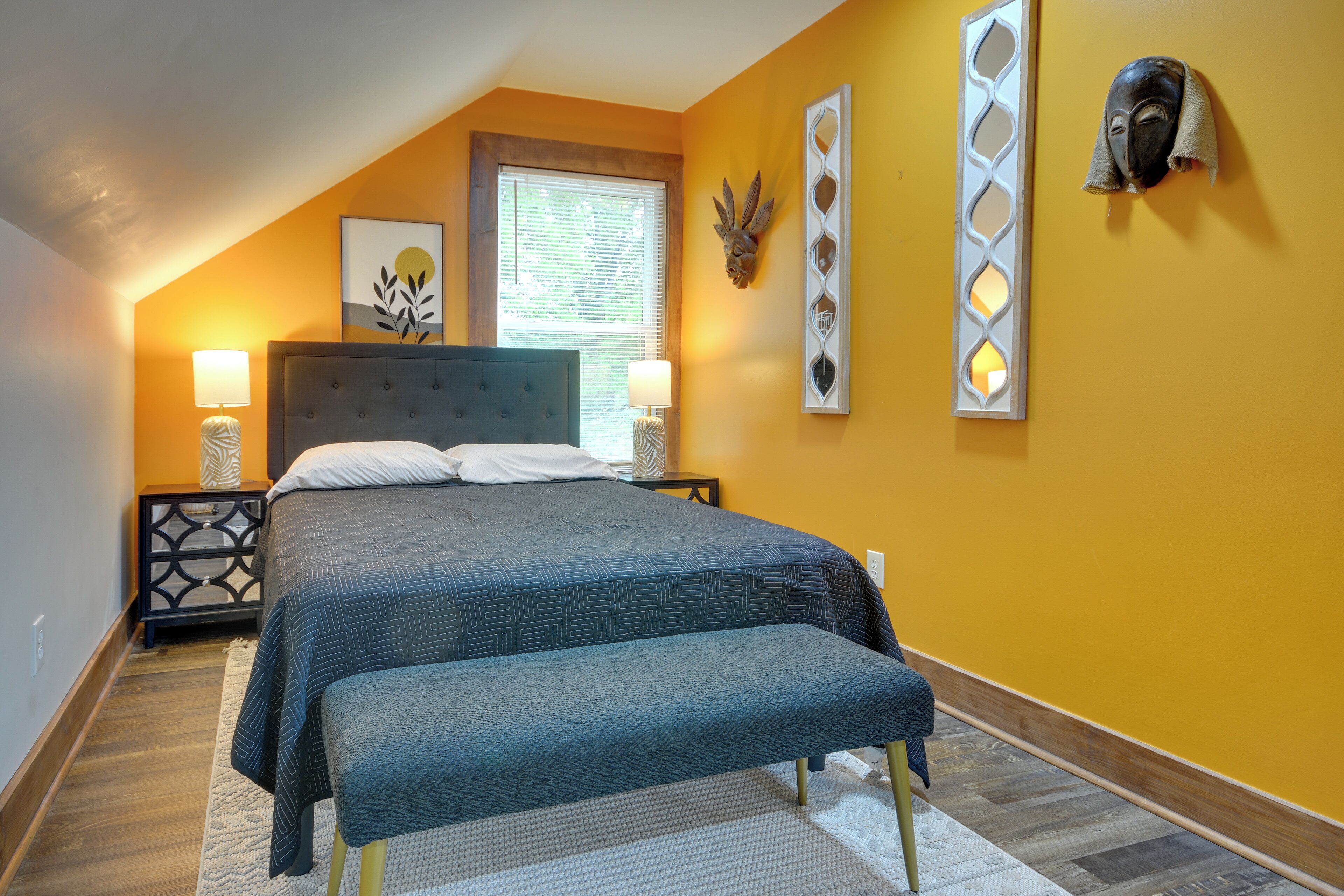 A bedroom with a bed, a bench, a nightstand, a wall-mounted headboard, a window with blinds, and a wall decoration.