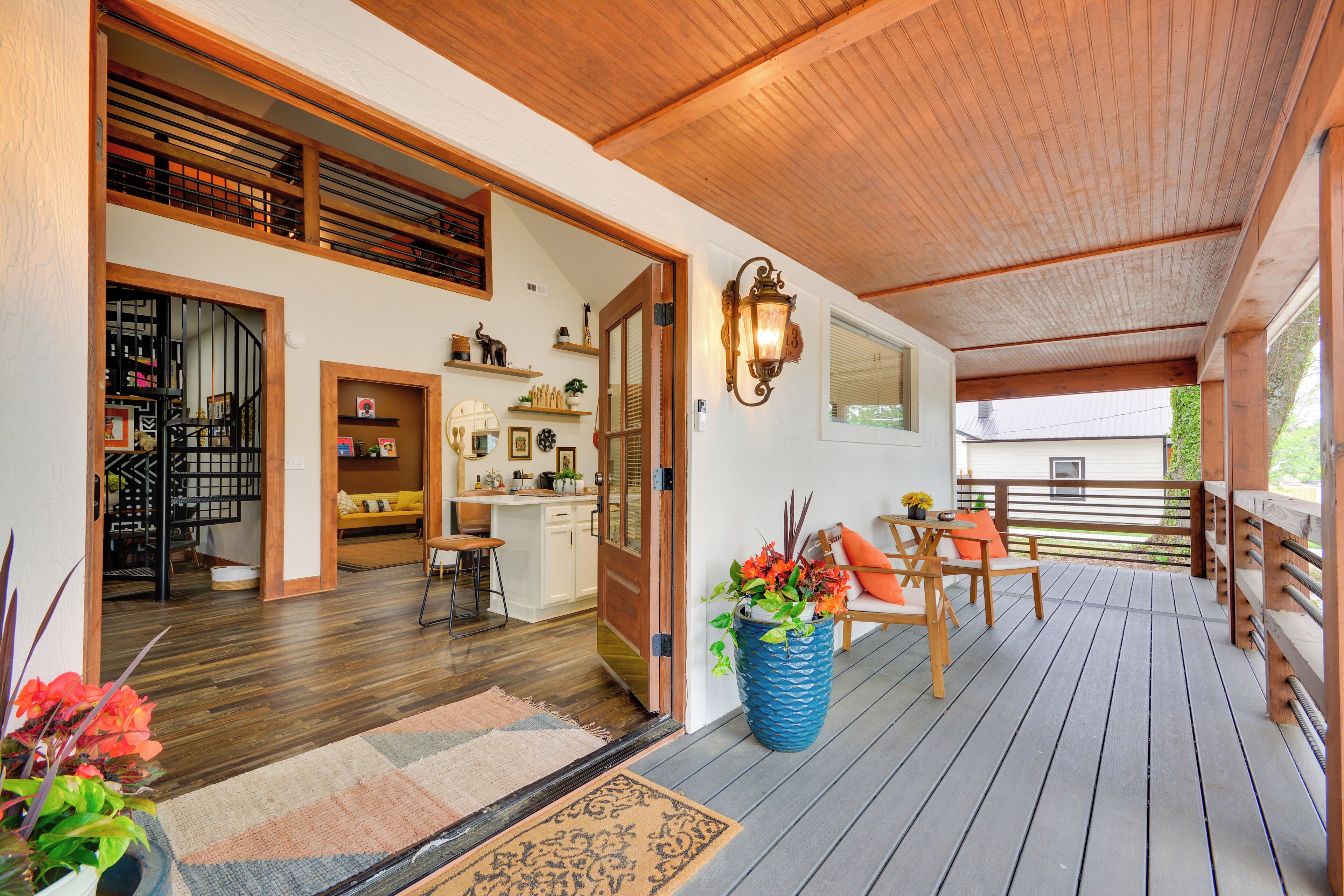 Um deck de madeira com área de estar externa, um vaso grande com planta e uma porta que dá para um espaço interior com cozinha e sala de jantar.