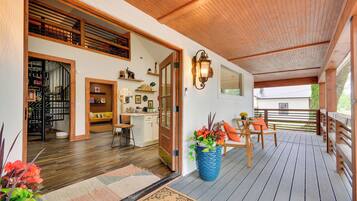 Um deck de madeira com área de estar externa, um vaso grande com planta e uma porta que dá para um espaço interior com cozinha e sala de jantar.