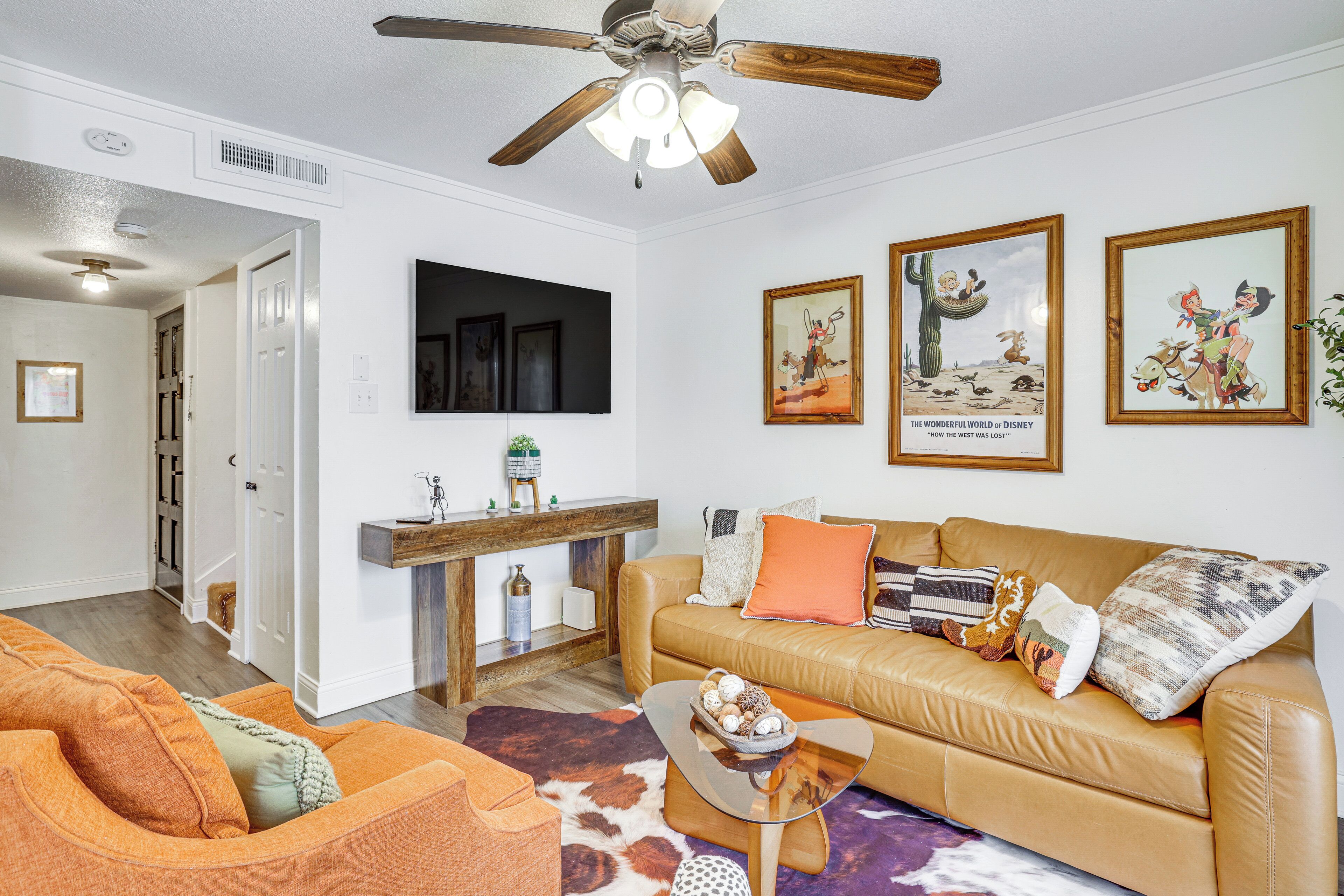 Un salon avec un canapé, une table basse et un ventilateur de plafond. Des tableaux encadrés ornent le mur, et une console en bois est également présente.