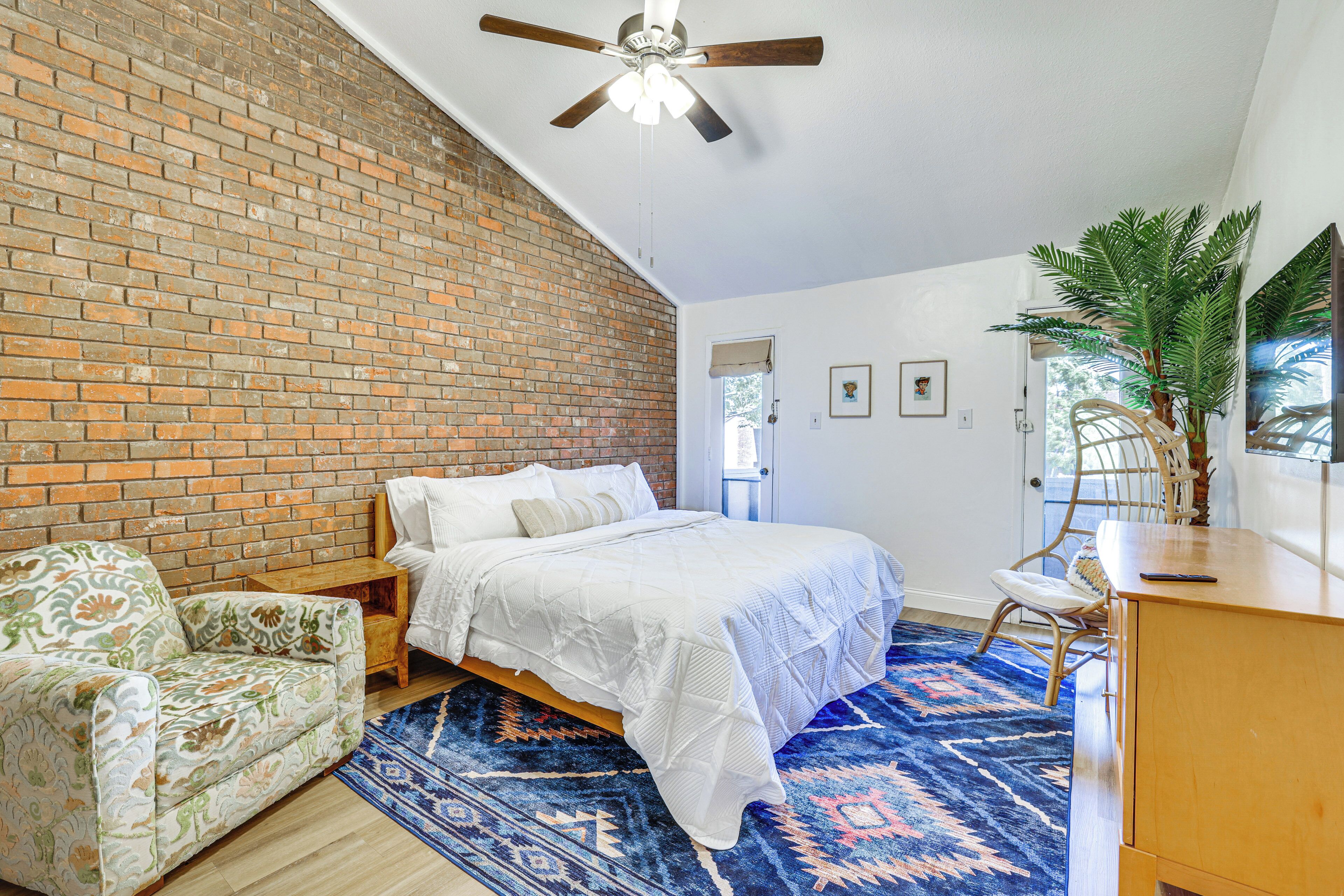 A bedroom with a brick wall, a bed with white bedding, a blue patterned rug, a ceiling fan, and a potted plant.