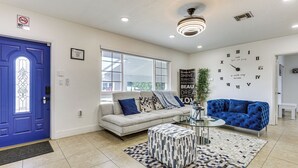 Una sala de estar con una puerta azul, un sofá, un sillón azul, un reposapiés estampado y un reloj de pared.