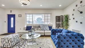 Una sala de estar con una puerta azul, un sofá, un reposapiés azul, una mesa de centro de vidrio y un reloj de pared.