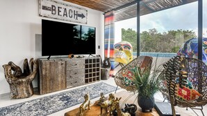 A living room with a television, wooden cabinet, decorative statues, a woven chair, and a view of the outdoors.